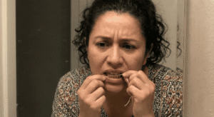Primer plano de una mujer mexicana con el ceño fruncido y una expresión de preocupación mientras usa hilo dental en sus dientes frontales en un baño.